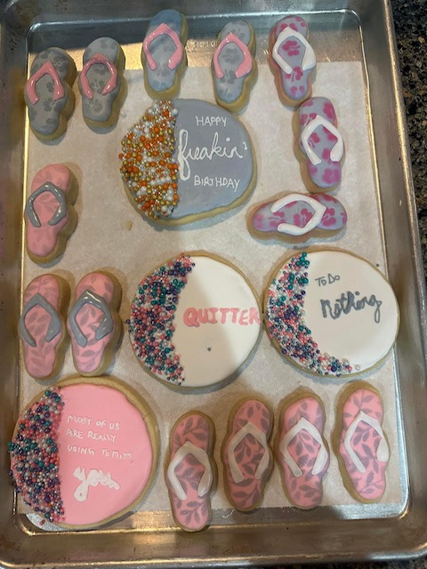 A baking tray filled with decorated cookies. Most are flip-flop shaped with pink and white icing. Round cookies include messages like "HAPPY freakin' BIRTHDAY," "QUITTER," "MOST OF US ARE REALLY GOING TO MISS YOU," and "todo NOTHING." Some have colorful sprinkles—perfect for a retirement party!