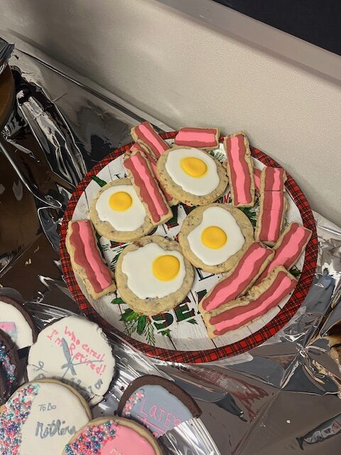 A plate of cookies, perfect for a retirement party, is decorated to look like bacon and eggs. The cookies are frosted with pink and white icing for the bacon and white icing with a yellow center for the eggs. The plate sits on a foil-covered surface alongside other festive cookies.