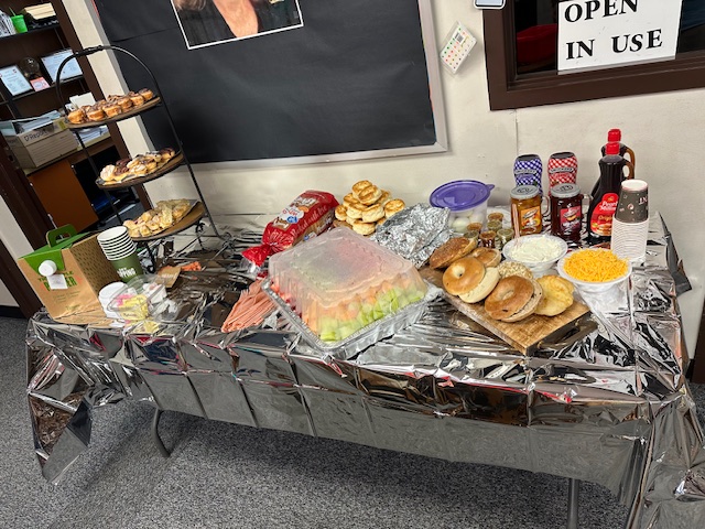 A table with a silver tablecloth displays a variety of breakfast foods, including bagels, croissants, spreadable toppings, a fruit tray, pastries, and syrup bottles. A tiered stand holds more pastries. The setup is in front of a wall adorned with an "OPEN IN USE" sign for the retirement party festivities.