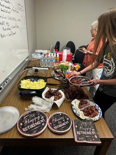 A person serves food from a table at a Retirement Party. The table is set with "Happy Retirement" and "Cheers to Retirement" plates, alongside dishes like scrambled eggs, bacon, and various vegetables. Another person is in the background, near the whiteboard.