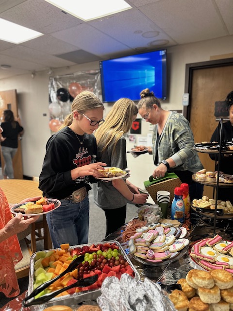 A group of people is gathered around a table filled with a variety of food, including fruit, pastries, and cookies. At the retirement party, two young girls in the foreground are holding plates and serving themselves while a woman stands behind them, assisting. A TV and decorations are in the background.