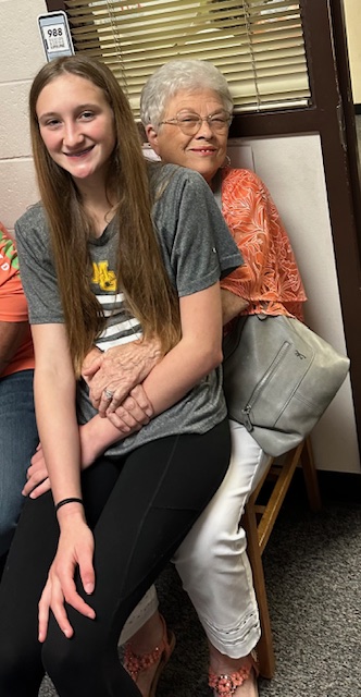 A young girl with long brown hair sits on an older woman's lap. The older woman, wearing glasses and a floral orange blouse, smiles and hugs the girl from behind. They are indoors at a retirement party, with part of another person visible beside them. There is a window in the background.