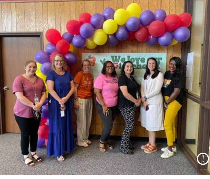 Seven people stand in a row, smiling at the camera. They are in front of a “Welcome to Your New School Year” banner decorated with a colorful balloon arch, lending the scene an air of celebration typically seen at a retirement party. The setting appears to be a school hallway or office area.