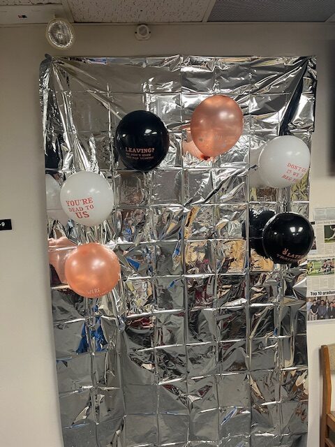 A wall decorated with a shiny silver foil backdrop sets the scene for the retirement party. Several balloons in white, black, and pink bear phrases like "YOU'RE THE TO MY BUS" and "DON'T BE A STRANGER." In the bottom right corner, scattered images and newspaper clippings add a nostalgic touch.