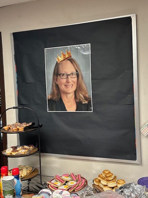 A bulletin board displays a photo of a woman wearing a digital crown at her Retirement Party. In front of the board, there is a table with various pastries and snacks, including cookies, biscuits, and a tiered tray with assorted treats.