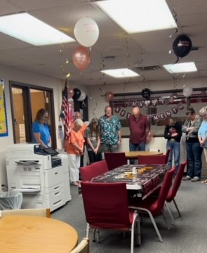 A group of people stand in a decorated room with balloons and a "Happy Retirement" banner, celebrating at the retirement party. They appear to be bowing their heads as if in prayer or reflection. A long table with decorations and chairs is in the center, and a printer is on the left side.