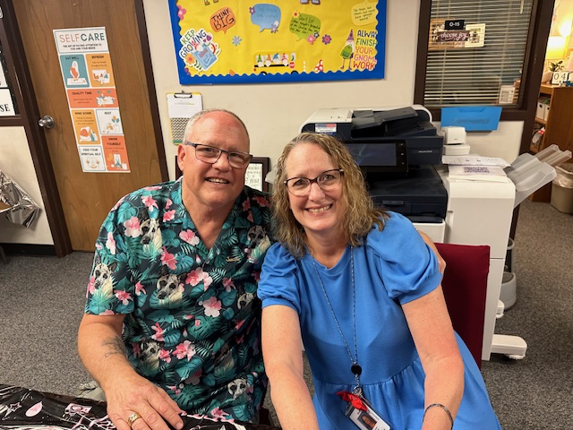 A man and a woman smile at the camera while seated at a table, celebrating a retirement party. The man wears glasses and a tropical shirt, and the woman wears glasses and a blue dress. Behind them is a bulletin board with colorful papers and a printer.